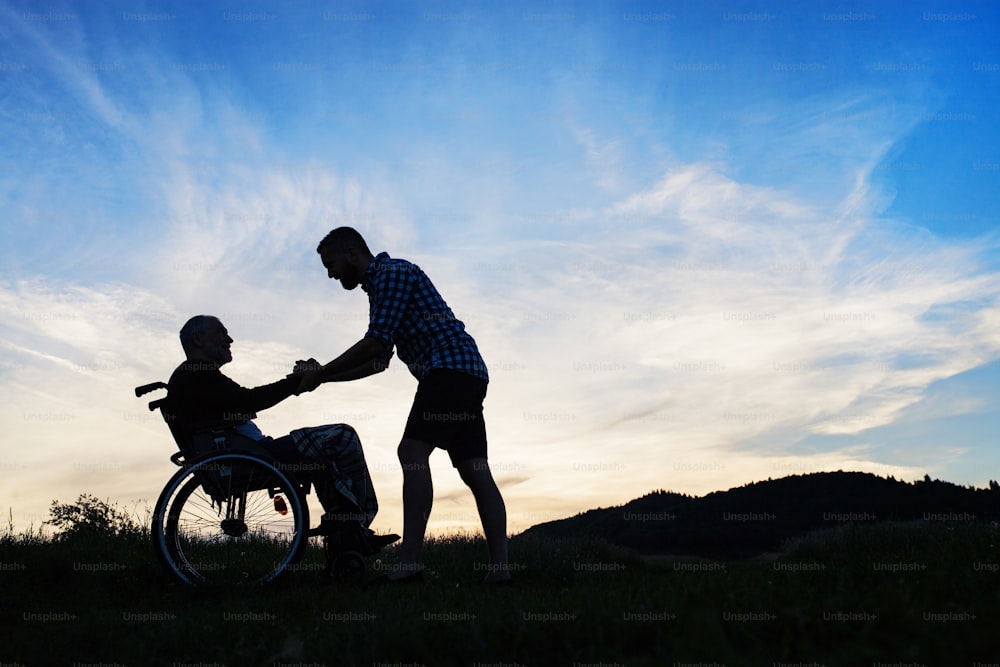 A silhouette of adult son looking at his senior father in wheelchair in nature at sunset. Copy space.