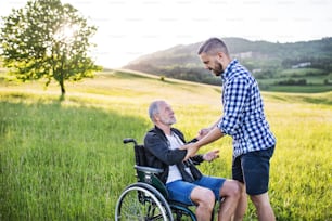 An adult hipster son looking at his senior father in wheelchair on a walk on a meadow in nature at sunset.