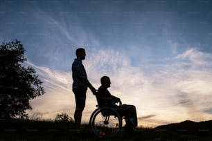 Un portrait en silhouette d’un fils adulte avec un père âgé en fauteuil roulant lors d’une promenade dans la nature au coucher du soleil.