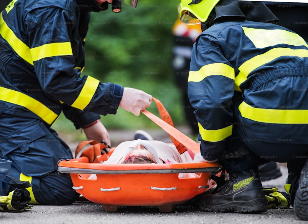 Unrecognizable firefighters putting a young injured woman into a plastic stretcher on the road after a car accident. Close up.