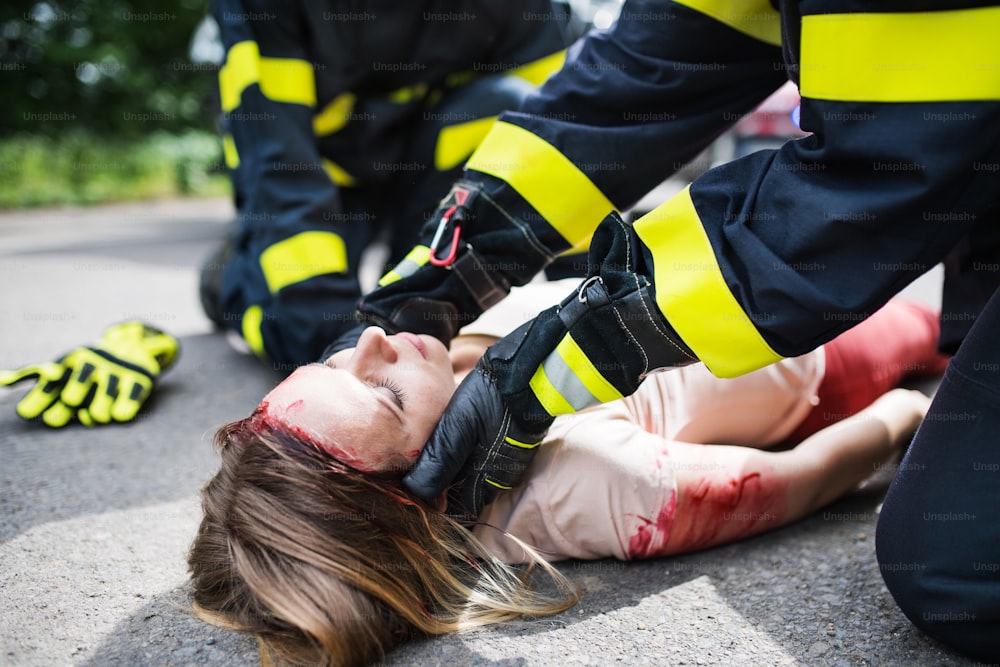 Bombeiros irreconhecíveis ajudando uma jovem ferida deitada na estrada após um acidente, inconsciente.