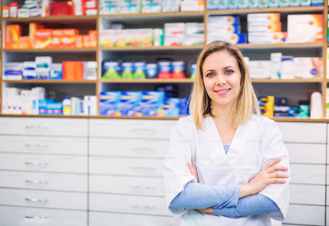 A young Asian female pharmacist in a white coat, arms crossed, in a pharmacy