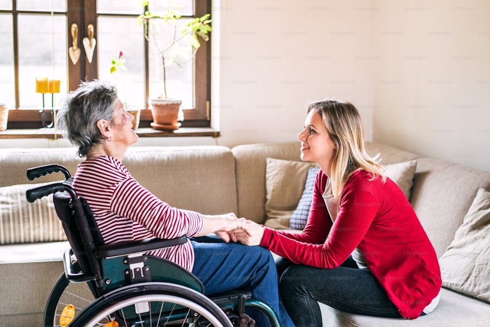 휠체어를 탄 할머니와 성인 손녀가 집 소파에 앉아 손을 잡고 있다.