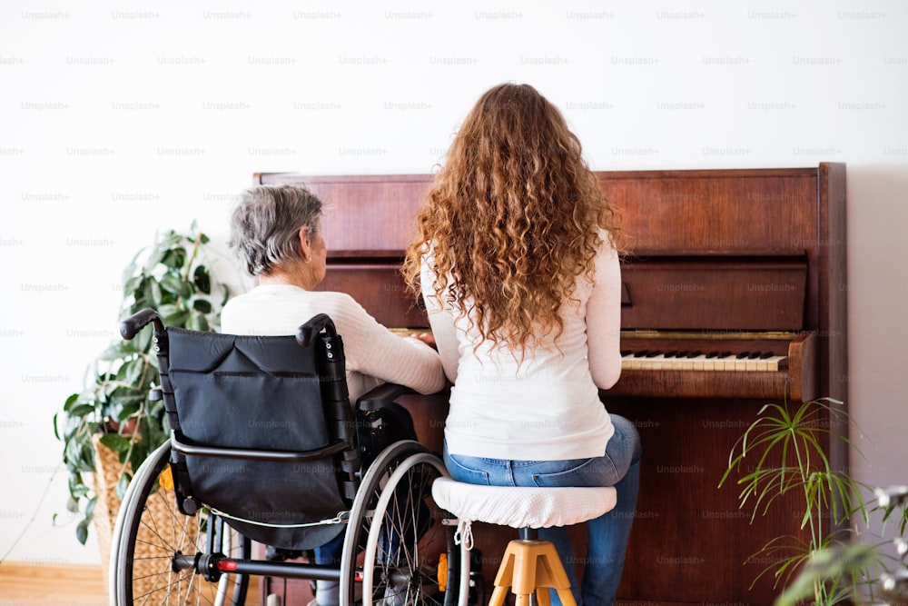 A teenage girl with grandmother in wheelchair playing the piano at home. Family and generations concept. Rear view.