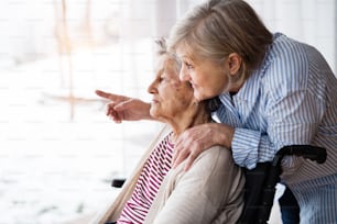 Eine ältere Mutter mit ihrer Tochter zu Hause, die aus dem Fenster schaut. Eine Pflegekraft, die einer behinderten älteren Frau im Rollstuhl hilft.