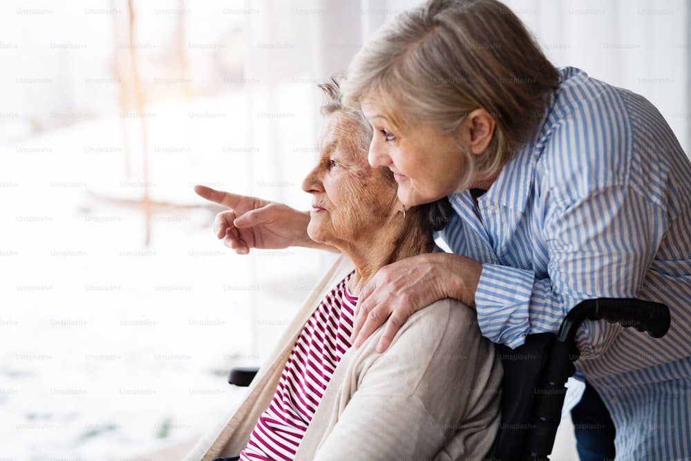 Uma mãe idosa com sua filha em casa olhando pela janela. Um cuidador que auxilia uma mulher idosa com deficiência em cadeira de rodas.