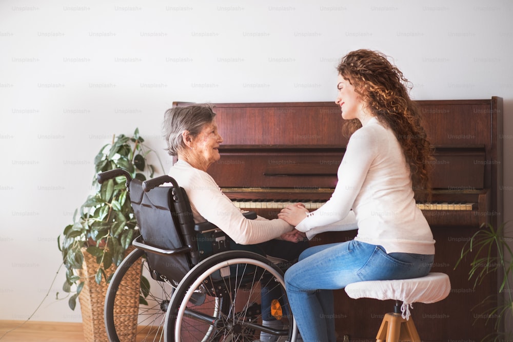 A teenage girl with grandmother in wheelchair sitting by the piano at home, holding hands.