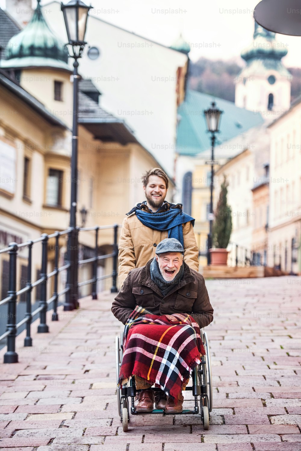 Père en fauteuil roulant et jeune fils en promenade, riant. Un soignant aidant un homme âgé handicapé.