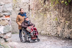 Father in wheelchair and young son on a walk in old town. A carer assisting disabled senior man.