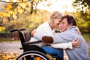 Senior couple in autumn nature. Man and woman in a wheelchair on a walk, hugging.