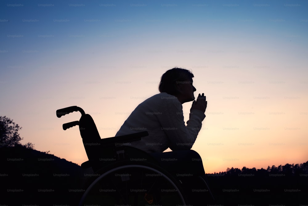 Eine Silhouette einer älteren Frau im Rollstuhl in der Natur am Abend. Eine Frau, die in der Abenddämmerung betet.