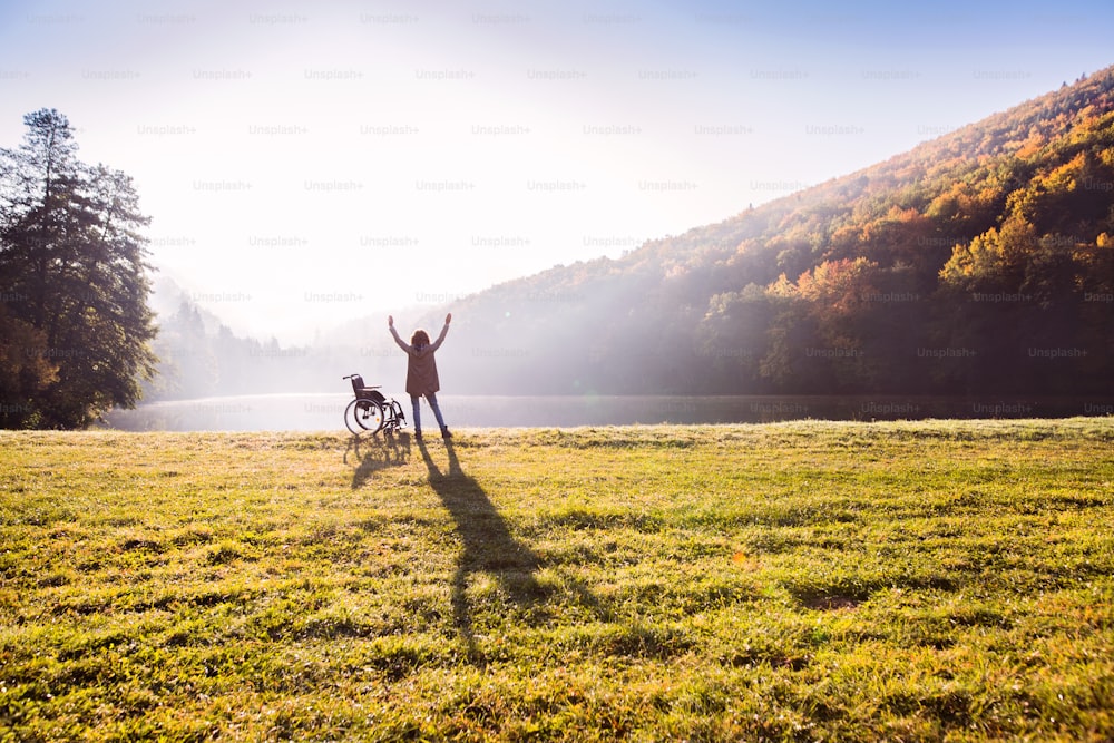 Mulher sênior em pé ao lado da cadeira de rodas em uma natureza de outono. Uma mulher relaxando à beira do lago no início da manhã. Vista traseira.