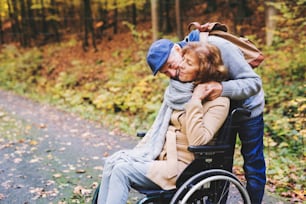 아름다운 가을 자연 속에서 산책하는 활동적인 노부부. 휠체어를 탄 남자와 여자가 숲 속을 걷고 있다.
