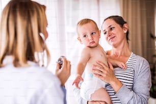 Young female doctor examining a baby with stethoscope in her office. Young mother holding a boy.