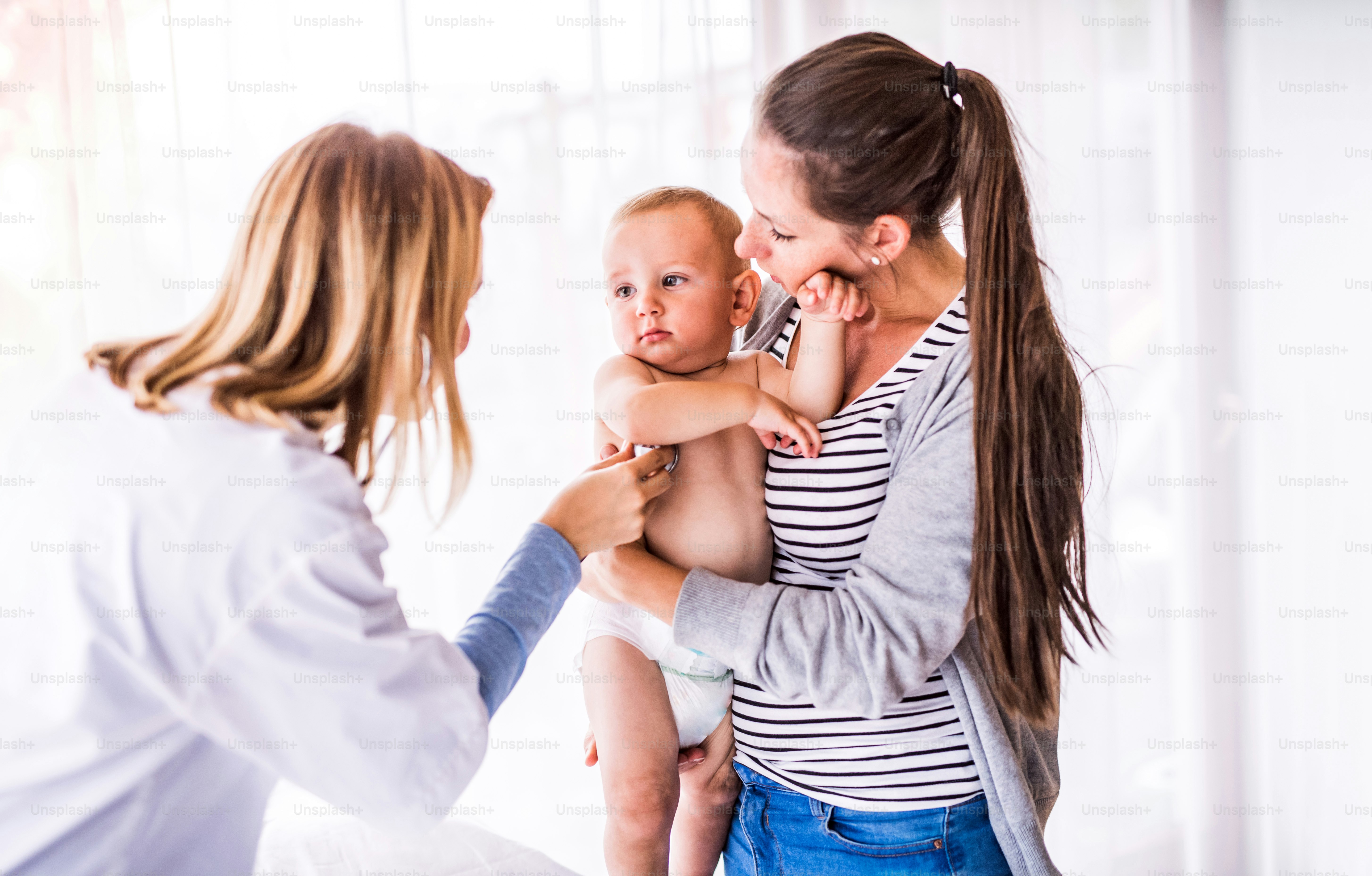 Young female doctor examining a baby with stethoscope in her office. Young mother holding a boy.