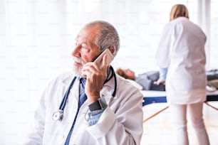 Médecin principal avec smartphone dans son bureau, faisant un appel téléphonique. Une infirmière et un patient couché à l’arrière-plan.