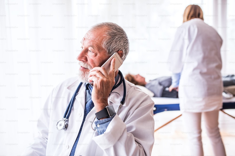 Médico senior con teléfono inteligente en su oficina, haciendo una llamada telefónica. Una enfermera y un paciente acostado en el fondo.