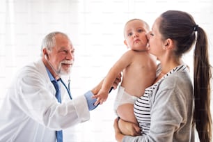 Senior male doctor examining a baby with stethoscope in his office. Young mother holding a boy.