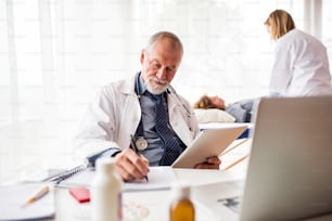 Senior doctor with tablet in his office. A nurse and a lying patient in the background.