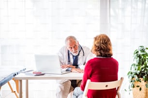 Männlicher Arzt mit Laptop im Gespräch mit einer älteren Frau in seinem Büro.