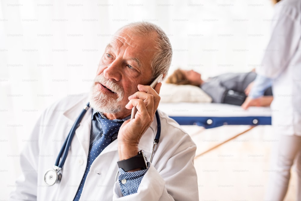 Médico senior con teléfono inteligente en su oficina, haciendo una llamada telefónica. Una enfermera y un paciente acostado en el fondo.