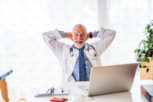 Senior male doctor with laptop at the office desk, sitting back in his chair with his hands behind his head.