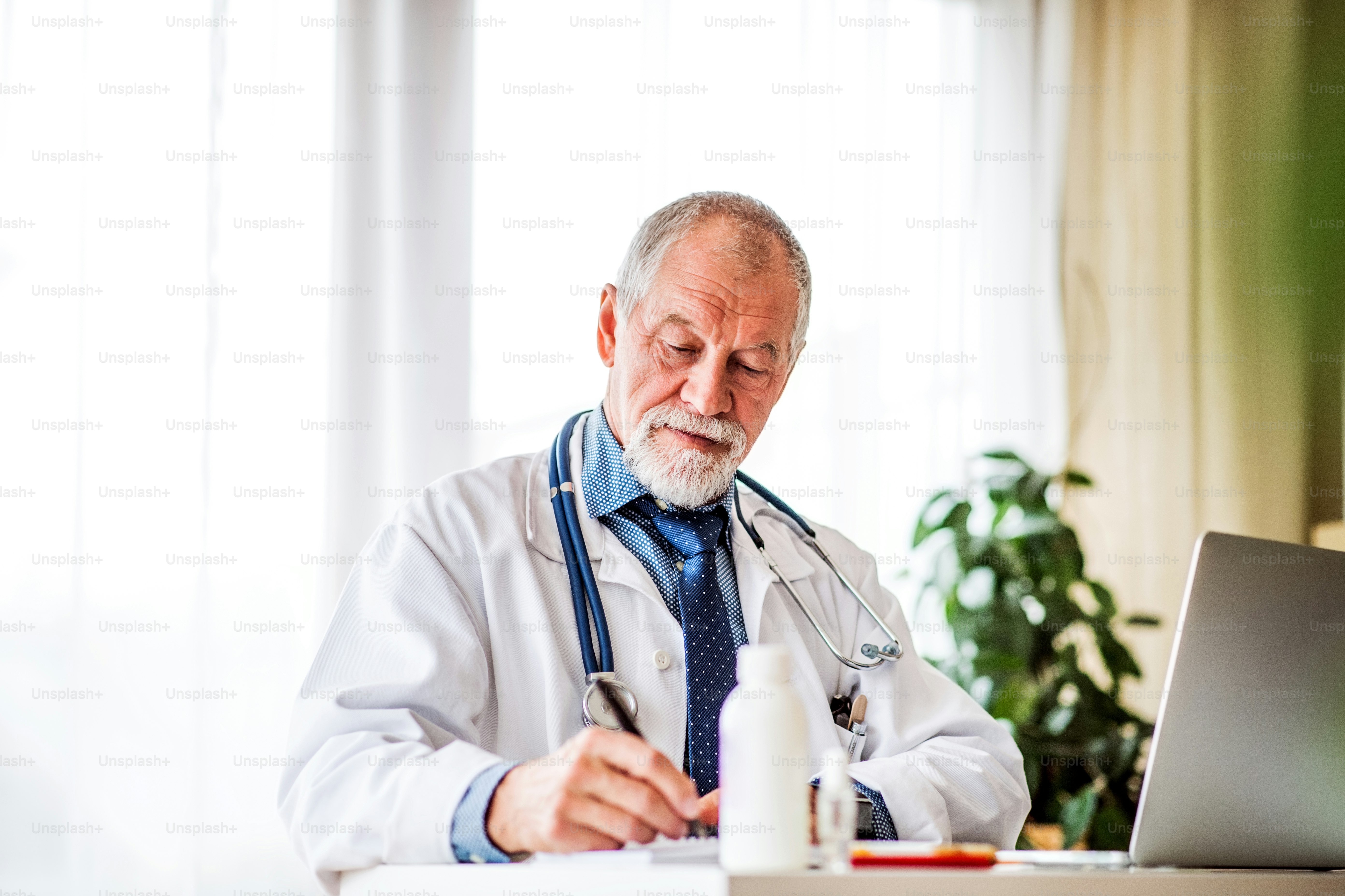 Médico masculino senior con computadora portátil en el escritorio de la oficina., tomando notas.