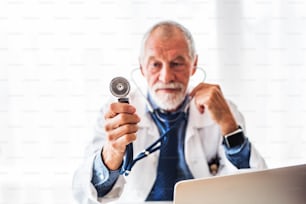 Leitender männlicher Arzt mit Laptop am Schreibtisch, der ein Stethoskop hält.