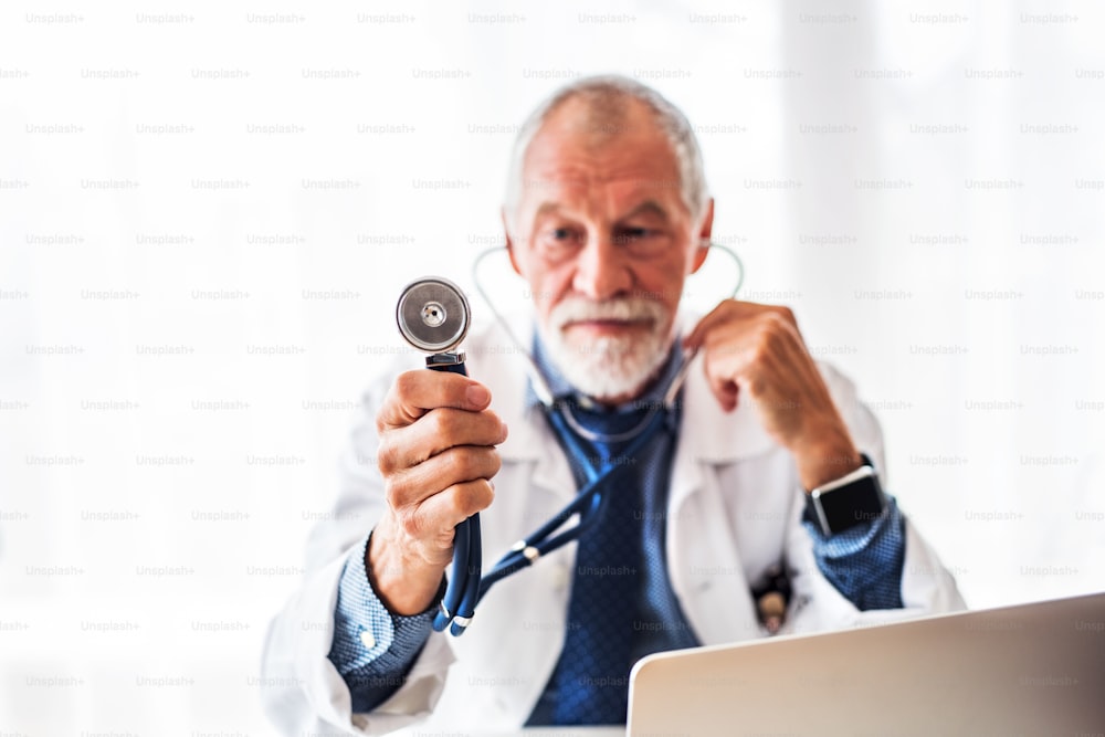 Senior male doctor with laptop at the office desk, holding a stethoscope.