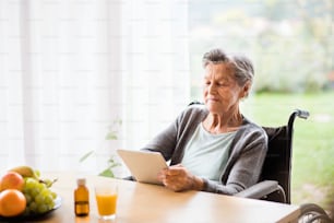 Donna anziana in sedia a rotelle con tablet a casa. Una donna anziana seduta al tavolo.