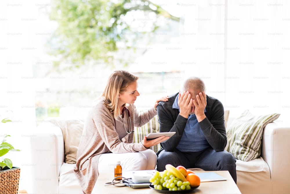 Health visitor and a senior man during home visit. A female nurse or a doctor showing test results on a tablet. Man feeling disappointed, covering face with hands.