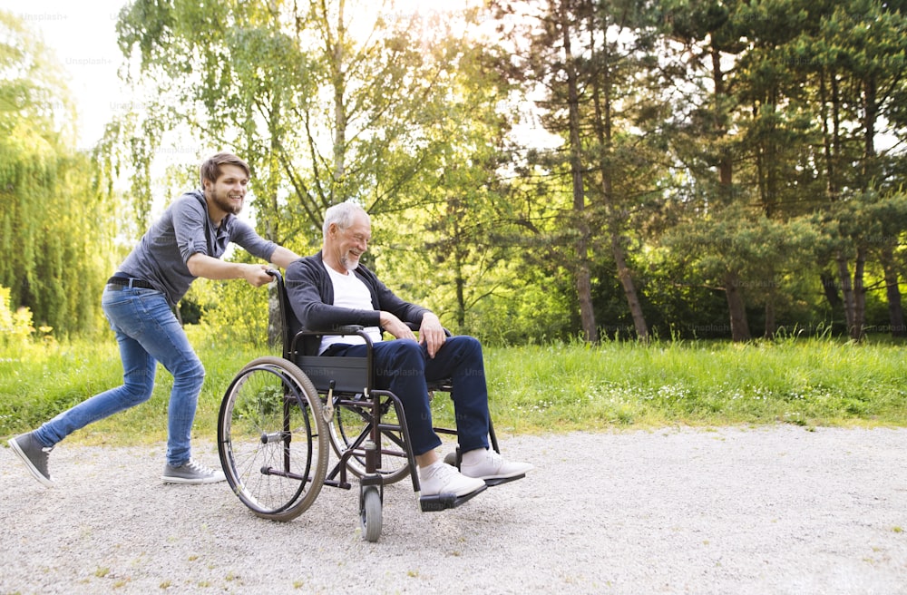 Giovane figlio hipster che corre con padre disabile in sedia a rotelle al parco, divertendosi insieme. Assistente che assiste l'anziano disabile.