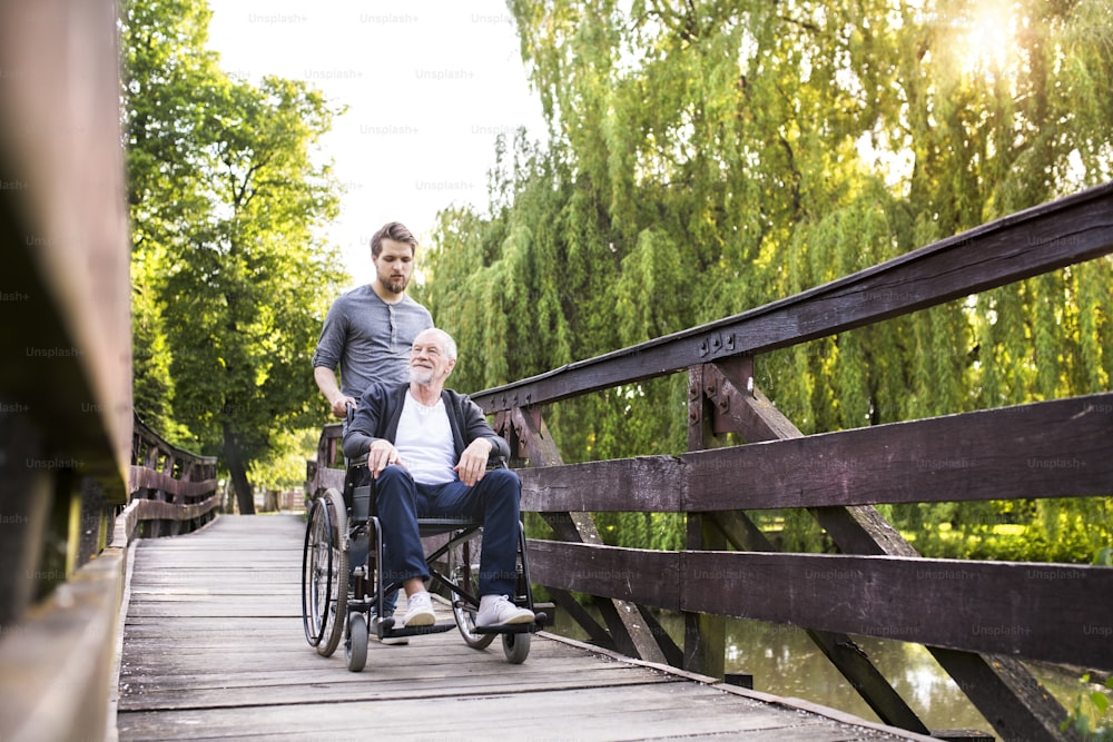 Young hipster son walking with disabled father in wheelchair on wooden bridge at park. Carer assisting disabled senior man.