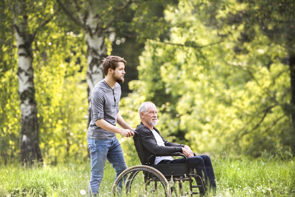 Giovane figlio hipster che cammina con il padre disabile in sedia a rotelle al parco. Assistente che assiste l'anziano disabile.