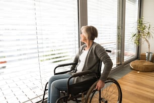 Mulher idosa deficiente em cadeira de rodas em casa em sua sala de estar, olhando pela janela.