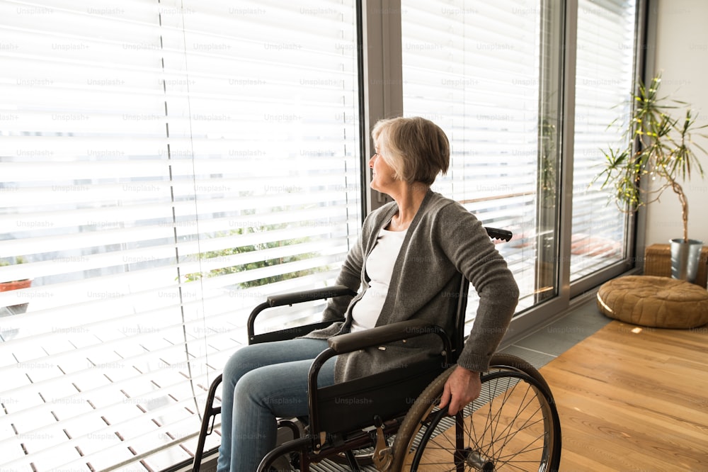 Donna anziana disabile in sedia a rotelle a casa nel suo soggiorno, guardando fuori dalla finestra.
