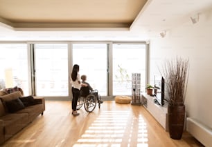 Mulher idosa deficiente em cadeira de rodas em casa em sua sala de estar, com sua filha pequena cuidando dela.