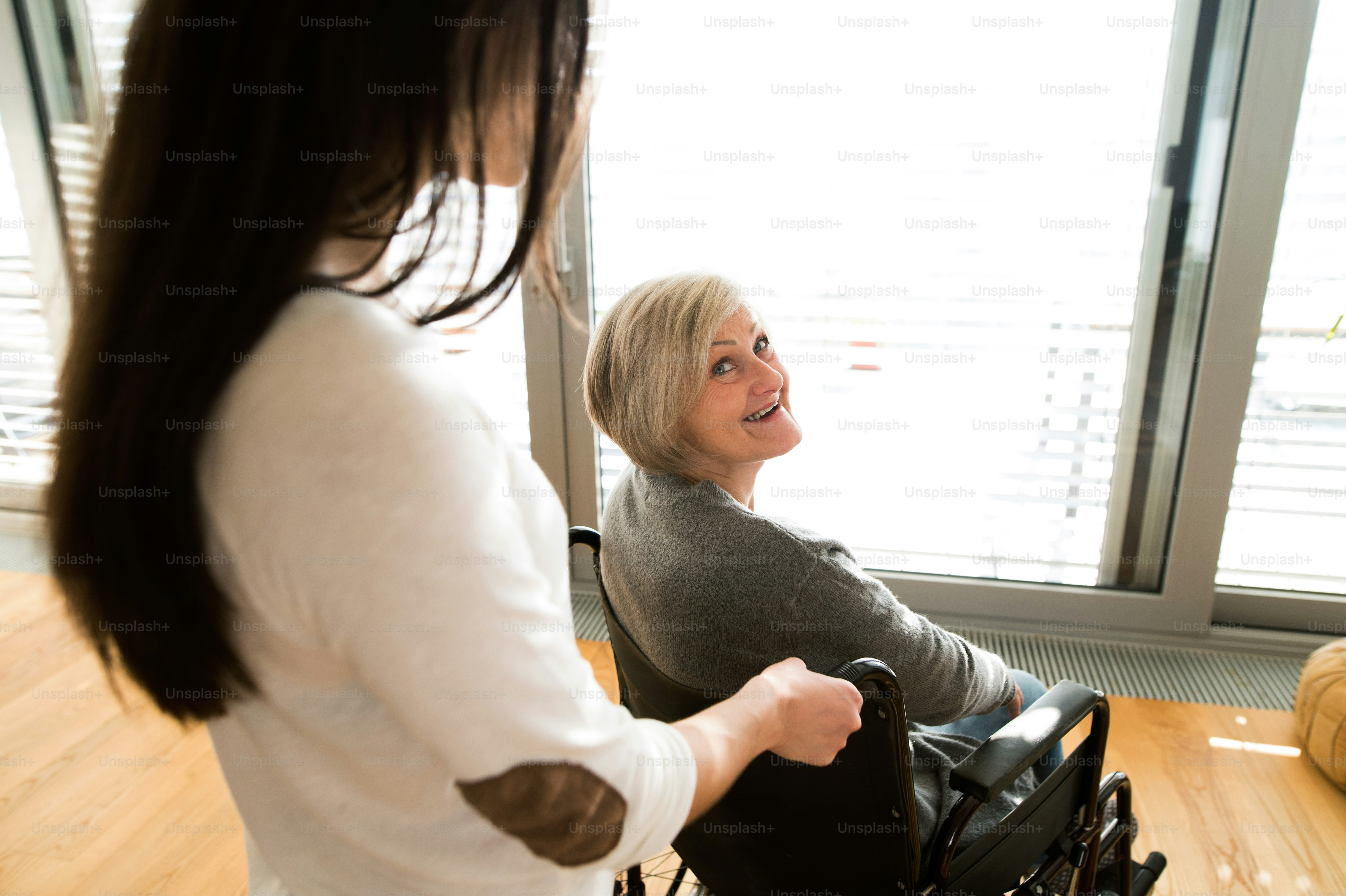 Disabled senior woman in wheelchair at home in her living room, with her young daughter caring for her.