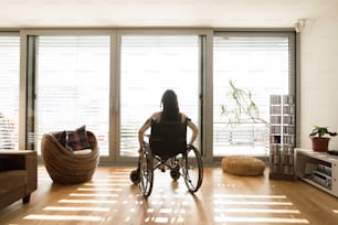 Bella giovane donna disabile in sedia a rotelle alla finestra di casa nel suo soggiorno. Veduta posteriore.