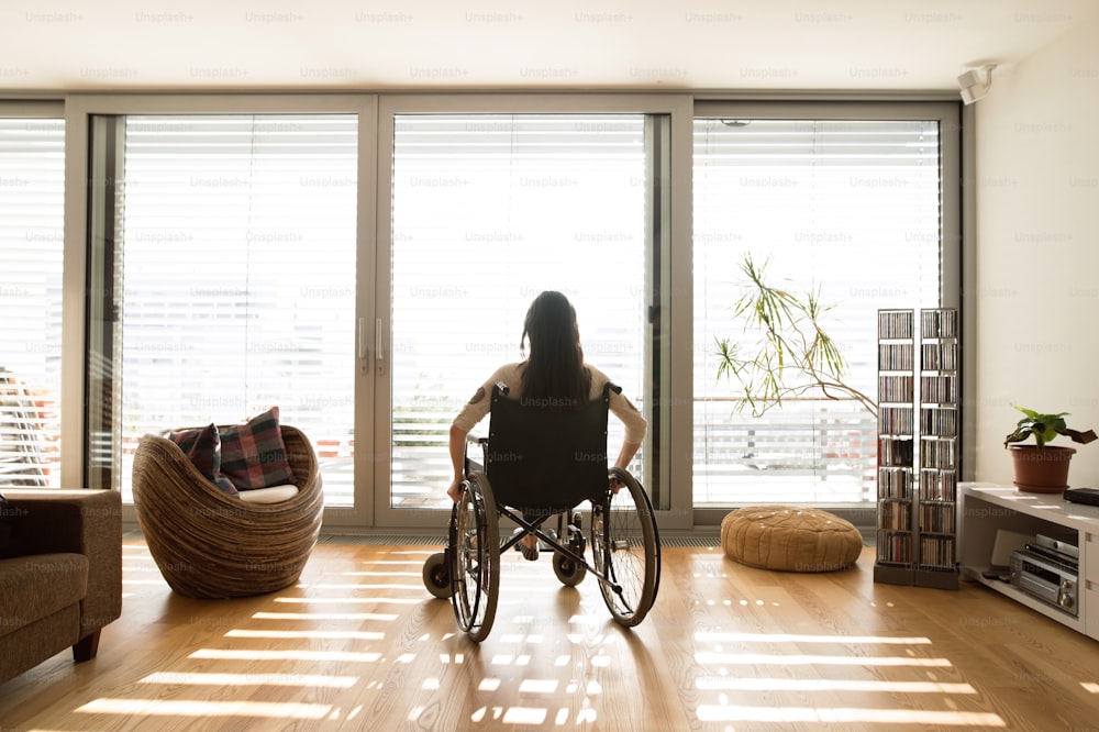 Mulher deficiente jovem bonita na cadeira de rodas na janela em casa em sua sala de estar. Vista traseira.