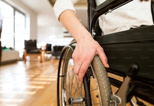 Unkenntliche junge behinderte Frau im Rollstuhl zu Hause in ihrem Wohnzimmer. Nahaufnahme ihres Arms, der auf dem Rad liegt.