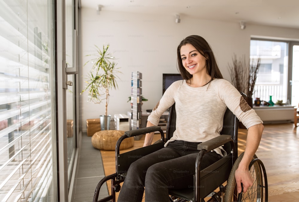 Mulher deficiente jovem bonita na cadeira de rodas na janela em casa em sua sala de estar