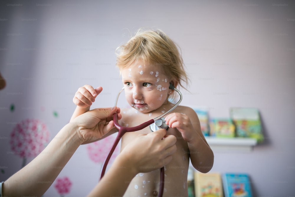 Menina de dois anos de idade em casa doente com varicela, creme antisséptico branco aplicado à erupção cutânea. Mãe dando seu estetoscópio.