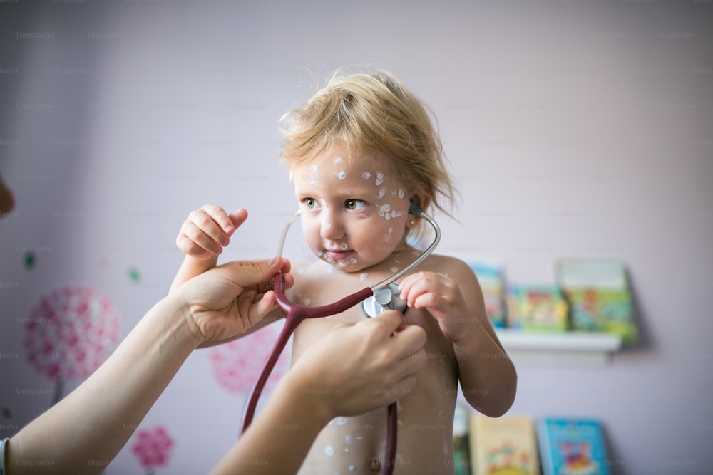 Little two year old girl at home sick with chickenpox, white antiseptic cream applied to the rash. Mother giving her stethoscope.