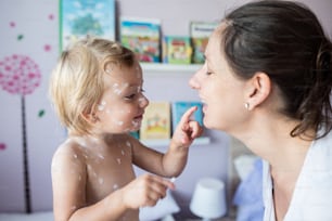 Niña de dos años en casa enferma de erupción de varicela, aplicando crema antiséptica blanca a su madre