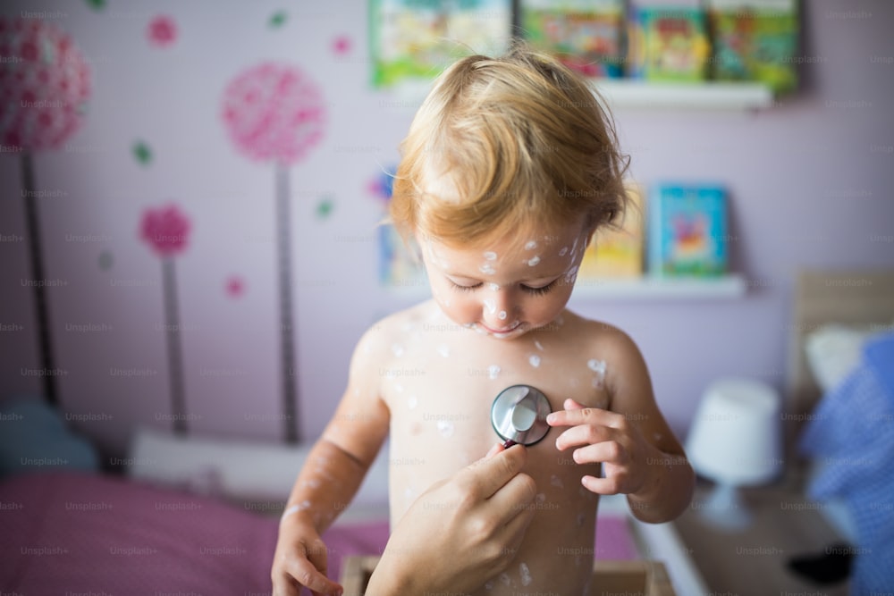 Kleines zweijähriges Mädchen zu Hause, das an Windpocken erkrankt ist, weiße antiseptische Creme, die auf den Hautausschlag aufgetragen wird. Spielt mit Stethoskop, nicht wiederzuerkennende Mutter hört auf ihren Herzschlag.