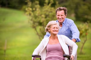 Senior man pushing woman sitting in wheelchair oustide in green autumn nature, laughing, runnning