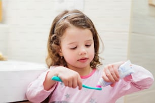 Linda niña en pijama en el baño poniendo una pasta de dientes en el cepillo de dientes