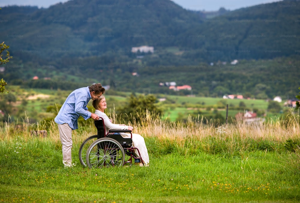 Senior man pushing woman sitting in wheelchair, kissing her, oustide in green autumn nature