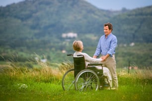 Homem sênior com mulher sentada na cadeira de rodas, de mãos dadas, oustide na natureza verde do outono, visão traseira